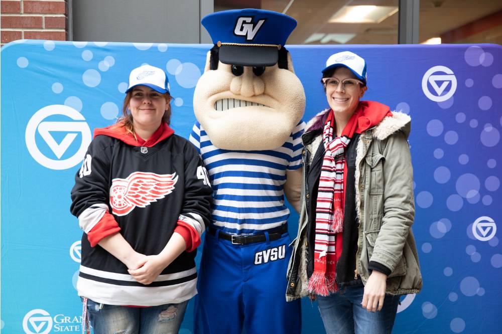 Two alumnae pose, wearing the Red Wings GVSU hat with Louie the Laker at the Detroit Red Wings GVSU Night